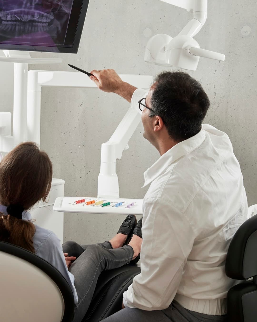 A dentist pointing at a panoramic x-ray on a wall monitor while a patient watches from the chair