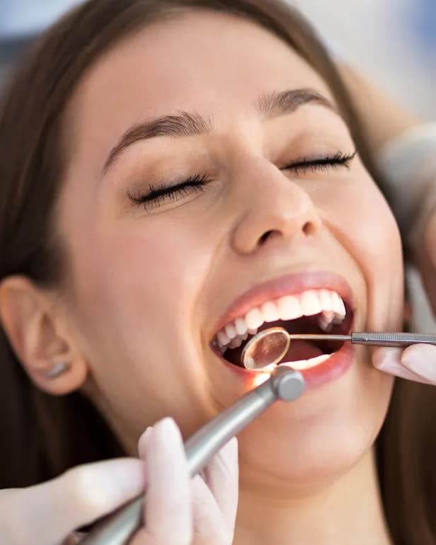 A smiling woman reclined in a dental chair while a dentist examines her teeth with a mouth mirror